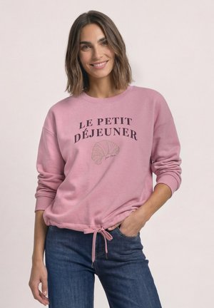 Femme souriante aux cheveux châtain moyen portant un sweat-shirt rose avec un motif "Le Petit Déjeuner" et croissant, associée à un jean bleu.