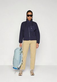 Tumesinine tõmblukuga jakk rinnataskuga, beežid püksid ja hele sinine seljakott. Modell kannab päikeseprille ja beeže tenniseid. Ühtlane sile tekstuur üle kogu rõivastuse.