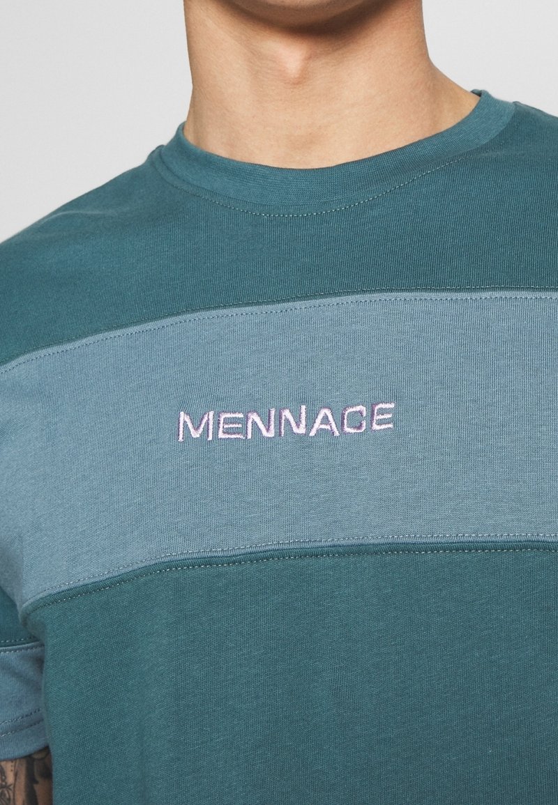 Petrolblå bomuld T-shirt med en lysere blå horisontal panel. Har lyserød broderet "MENACE" tekst på brystet. Rund hals og korte ærmer.