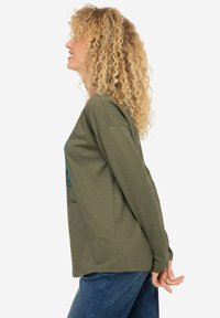 Koszula z długim rękawem w oliwkowym kolorze o luźnym kroju, z nadrukiem graficznym na przodzie. Materiał wydaje się miękki i lekki.