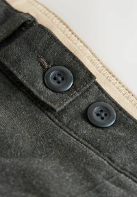 Detailní záběr tmavě šedé látky se dvěma černými čtyřdírovými knoflíky a béžovým zigzagovým stehem podél okraje.