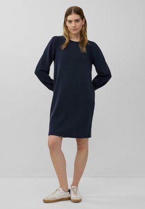Marineblaues Kleid mit langen Ärmeln, lässiger Passform, Rundhalsausschnitt, glattem Stoff und seitlichen Schlitzdetails, kombiniert mit weißen Sneakers.