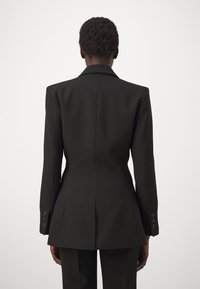 Blazer nero con una vestibilità su misura, spalle strutturate e bottoni decorativi sulle maniche. Texture liscia con una spacco sul retro.