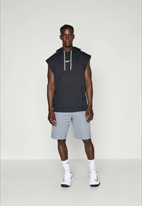 Hoodie senza maniche nera con logo Nike bianco, abbinato a pantaloni corti grigi chiari. L'outfit presenta una tasca frontale e un cappuccio regolabile con cordini.