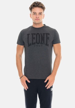 Jonge man met kort haar, draagt een donkergrijs "LEONE 1947" T-shirt en zwarte broek, staat met ontspannen handen voor zich.