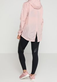 Chubasquero rosa claro con capucha, con puños elásticos y un diseño de espalda partida. Leggings negros con detalles texturizados. Zapatillas rosas.