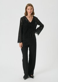 Sort blonde-dragt med lange ærmer, V-udskæring og dekorativ bindebånd foran. Har vide benåbninger og indviklet blonde detaljer.