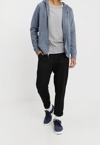 Modrá mikina na zip s dvěma kapsami přes šedé tričko, doplněná černými zúženými kalhotami a námořnickými teniskami. Jednoduchý, ležérní vzhled.