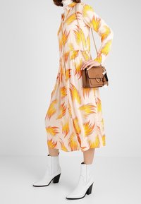 Abito a fantasia floreale in pesca con motivi di foglie gialle e arancioni, vita stretta, abbinato a stivali alla caviglia bianchi e una borsa a tracolla color beige.