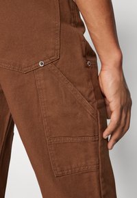 Brown fabric cargo pants featuring a side pocket, stitched accents, and silver-tone hardware details. Texture is smooth with a slight sheen.