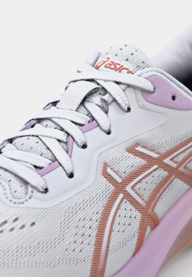 Des chaussures de sport blanches avec une tige en maille texturée, présentant des accents lilas et orange, des lacets plats et un col rembourré.