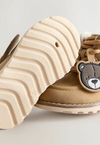 Kindersandfarbene Winterstiefel mit strukturiertem Boden und bärenförmigem Charme, der an sandfarbenen Schnürsenkeln befestigt ist, vor einem einfarbigen Hintergrund.