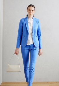 Světle modrý přizpůsobený blazer a odpovídající kalhoty, s bílým topem. Modelka nese béžovou kabelku a stojí na dřevěné podlaze před hladkou zdí.