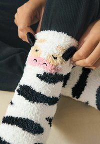 Vita fluffiga strumpor med svarta ränder och en koansikte-design, med strukturerat material och mjuka detaljer runt öronen och nosen.
