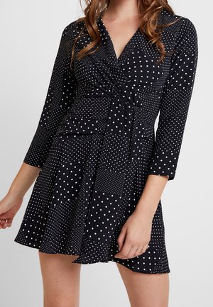 Frau, die ein schwarzes Wickelkleid mit weißen Punkten, Dreiviertelärmeln und ausgestelltem Rock vor hellem, einfarbigem Hintergrund trägt.