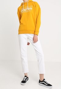 Osoba nosi musztardową żółtą bluzę z tekstem, białe spodnie dżinsowe 3/4 oraz czarne niskie trampki z białymi sznurówkami i podeszwami.