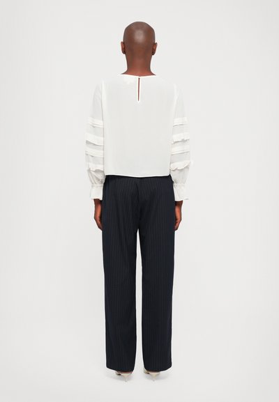 Personne avec la tête rasée portant une blouse blanche à manches longues avec des détails en ruffles et un pantalon large à rayures noires, vue de dos.