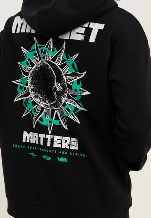 Sort hættetrøje med en grafik af en tekstureret central figur omgivet af pigge og grønne flammer samt tekst, der lyder "Matters" og "Shape your thoughts."