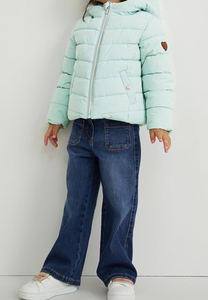 Niño con chaqueta acolchada azul claro, pantalones vaqueros azul oscuro y zapatillas blancas, de pie contra una pared lisa y clara.