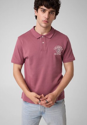 Junger Mann mit lockigem dunklem Haar, der ein mauvefarbenes Poloshirt mit weißem "California Guess Jeans"-Logo und hellblauen Jeans trägt, stehend vor einem schlichten Hintergrund.