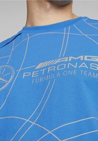 Blå bomullströja med en geometrisk design i vita linjer och logotyper. Inkluderar tryckt text: "AMG PETRONAS FORMULA ONE TEAM."