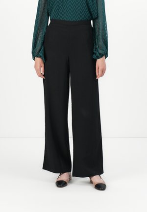Person med svarte vide bukser, en teal langermet topp med chevronmønster, og to-tonet beige og svarte flate sko, som står på hvitt gulv.