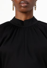 Black collared blouse with gathered detailing at shoulders, featuring subtle branding on collar and textured fabric. Gold chain earrings visible.
