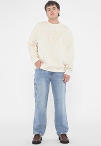 Crème sweatershirt met een driehoeklogo en vraagteken, gecombineerd met lichtblauwe cargobroek met zijzakken en een relaxte pasvorm. Brown schoenen.