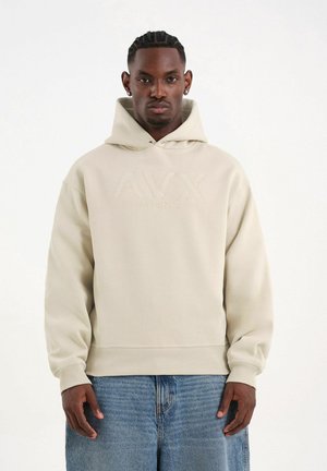 Man met ingevlochten haar die een beige AVIREX-hoodie en blauwe spijkerbroek draagt, staand tegen een effen witte achtergrond.