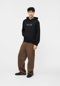 Νέος άνδρας που φοράει μαύρο φούτερ με κουκούλα και φαρδιά καφέ παντελόνια, στέκεται με το ένα χέρι στην τσέπη, σε απλό φόντο.