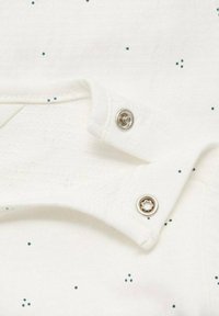 Tissu blanc avec de petits pois teal, présentant une fermeture à pression et un col texturé. Le design est simple et pratique.
