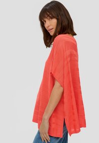 Koraalroze pullover met ruime mouwen, textuurstrepen, losse pasvorm en zijsplitten. Gecombineerd met blauwe jeans. Lichte stof.