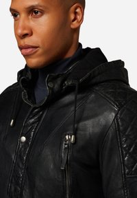 Schwarze Lederjacke mit Kapuze, ausgestattet mit gesteppten Akzenten, Druckknöpfen und einer mit Reißverschluss gesicherten Brusttasche. Texturierte Oberfläche und Kordelzugdetail.