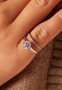 Three rings on a finger: a silver band with a large clear stone, a thin silver band with small clear gemstones, both with a polished finish.