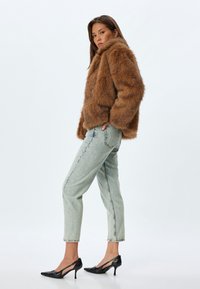 Ruskea tekoturkki takki, korkea kaulus, rento malli. Vaaleansiniset farkut käärittyine lahkeineen. Mustat lakatut nahkabootsit, joissa on terävä kärki ja leikattu muotoilu.