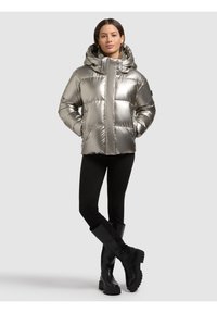 Giacca invernale argentata e imbottita con cappuccio, zip frontale e tasche laterali. Il materiale ha una finitura lucida e riflettente. Abbinata a pantaloni neri e stivali.