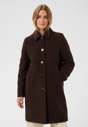 Femme portant un manteau marron foncé boutonné avec col, debout avec une main dans la poche devant un fond clair uni.