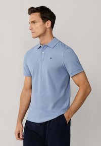 Mężczyzna ubrany w jasnoniebieską koszulkę polo z krótkim rękawem i małym wyszywanym logo, stoi z jedną ręką w kieszeni na jednolitym tle.