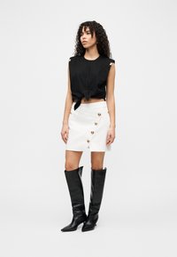 Μαύρο κοντό τοπ με λεπτομέρεια δεσίματος, σε συνδυασμό με λευκή φούστα τύπου wrap με χρυσά κουμπιά και μαύρα μπότες μέχρι το γόνατο.