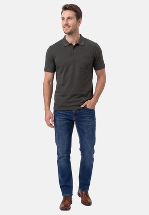 Mężczyzna w ciemnoszarej koszulce polo, niebieskich dżinsach i brązowych skórzanych butach, stojący z jedną ręką w kieszeni, uśmiechający się i patrzący w prawo.