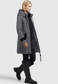 Grå quiltet parka med hætte, med sorte detaljer og store sidelommer. Bæret med sorte leggings og knæhøje sorte støvler.