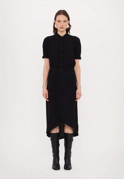 Robe noire avec un col de chemise à boutons, de courtes manches bouffantes et un ourlet asymétrique. Portée avec des bottes noires hautes. Texture lisse.