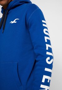 Blue hoodie made of soft fabric with a front kangaroo pocket, featuring a white logo and large text along the sleeve. Adjustable drawstring hood.