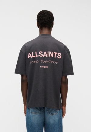 Oseba nosi temnosivo majico s kratkimi rokavi s pisanimi roza napisi "ALLSAINTS", japonskimi znaki in napisom "LONDON" na hrbtni strani, v kombinaciji z modrimi kavbojkami.