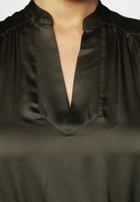 Blusa in satin verde scuro con scollatura a V, caratterizzata da morbidi plissettamenti sulle spalle e una texture liscia e lucida.