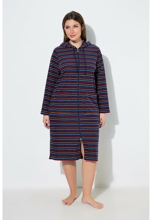 Frau steht barfuß und trägt einen knielangen, marineblauen gestreiften Bademantel mit Reißverschluss, Kapuze und Vordertaschen.