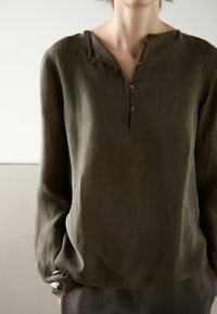 Chemise à manches longues vert olive foncé avec un col henley boutonné. Fabriquée dans un tissu texturé, présentant une légère brillance.