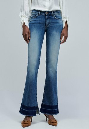 Pantalones vaqueros de mezclilla azul acampanados con una cintura de altura media, acabado deslavado y dobladillo de mezclilla oscura contrastante. Presentan un ligero deshilachado en el dobladillo.
