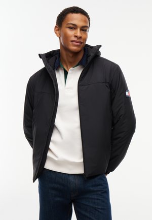 Jeune homme portant une doudoune noire par-dessus une chemise blanche à col et un jean foncé, debout devant un fond blanc uni.