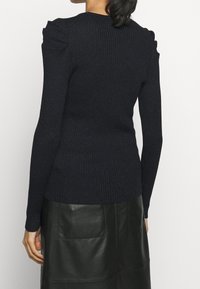 Eine Frau trägt einen schwarzen gerippten Langarmpullover mit gepufften Schultern und einen schwarzen Lederrock, von hinten gezeigt.